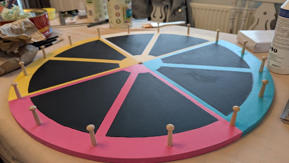 Wooden disk, yellow, white, cyan, magenta. Pegs stick up around the circumference. On a table with glue, paints etc. 