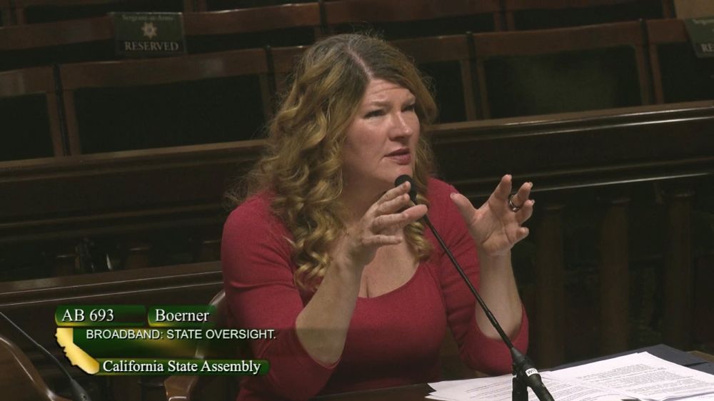 Assemblymember Boerner answers a question in Assembly Communications & Conveyance. At the bottom is a green logo with white font with the words "AB 693 Boerner Broadband State Oversight California State Assembly" with a gold logo of the State of California.