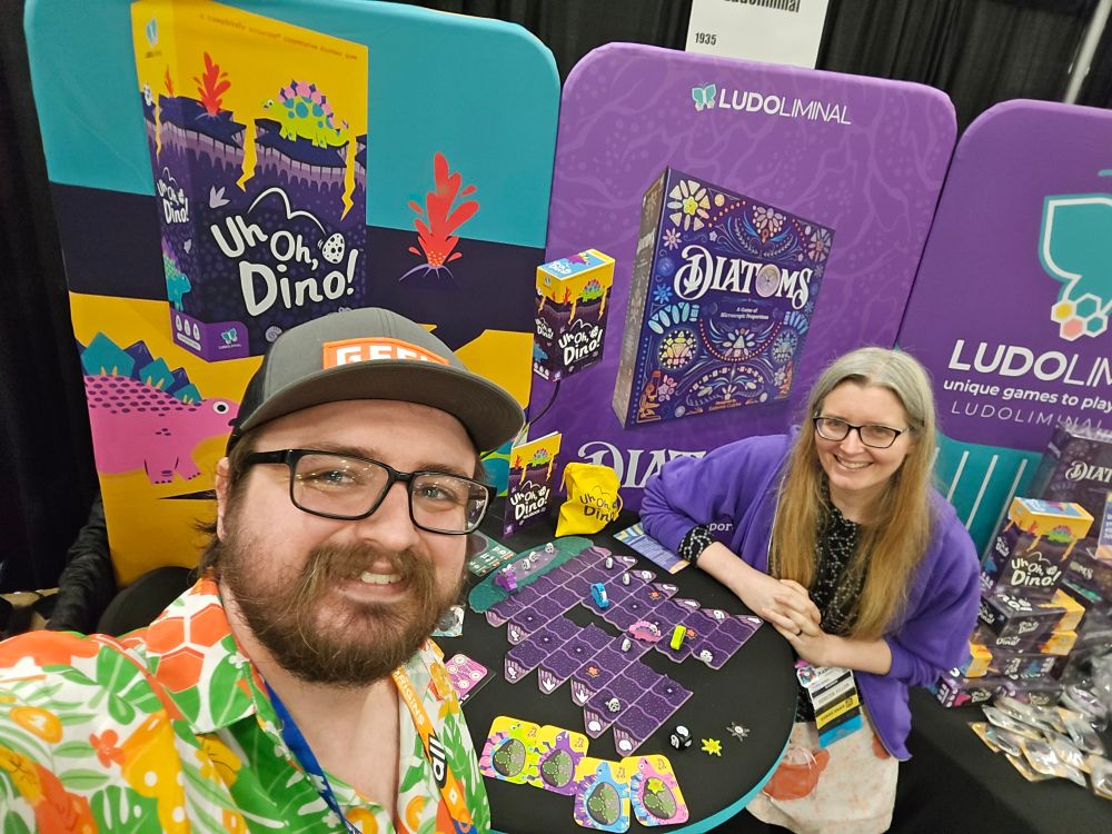 A very handsome man and an incredibly talented designer stand in front of a variety of vanders showcasing off Ludoliminal board games.