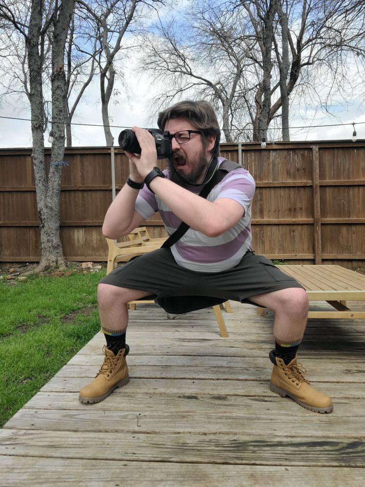 A very handsome man squats aggressively with a camera dressed in a nearly complete cosplay of Todd Snap.