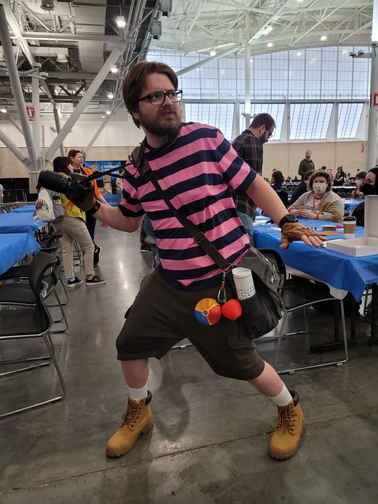 A very handsome man poses dramatically with a camera in hand, dressed in a full Todd Snap cosplay.