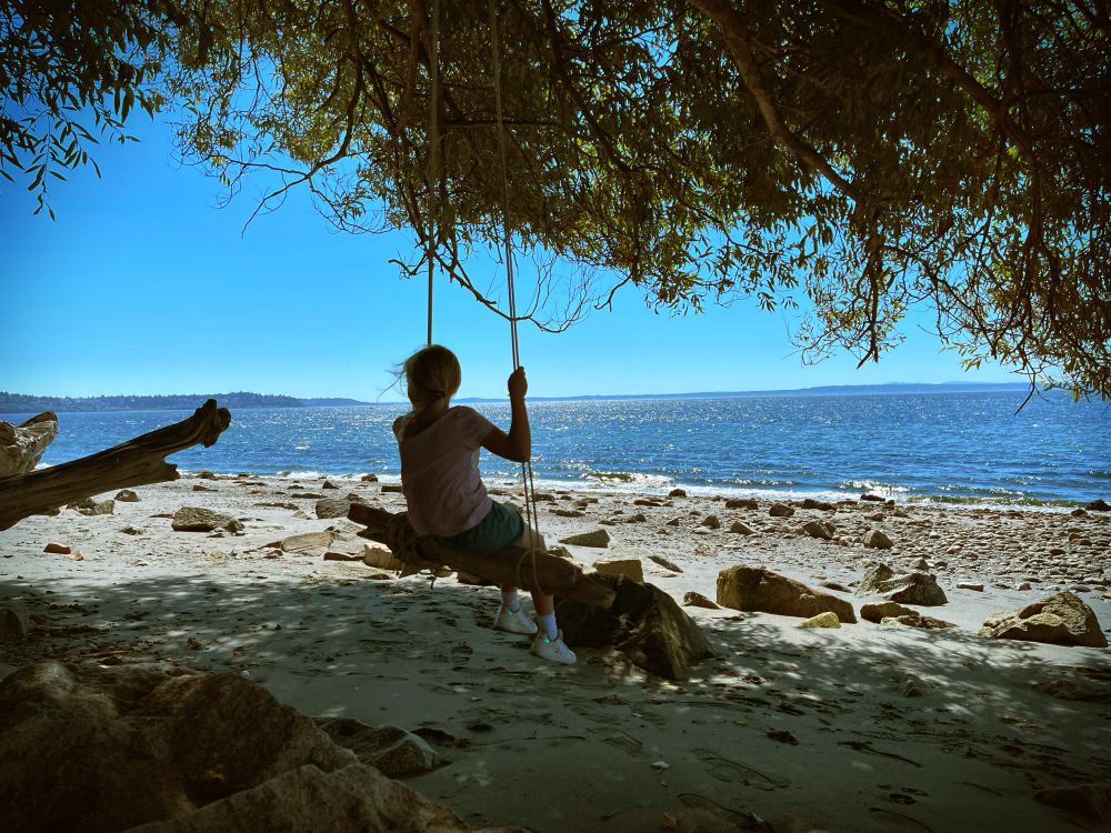 A young girl sits on a makeshift swing made of driftwood and found rope. The swing is on the beach of the Puget Sound. The girl is shadowed by the tree above her and is cast in silhouette against the bright blue water of the sound and rocks and sand of the beach. It’s a beautiful Summer day.