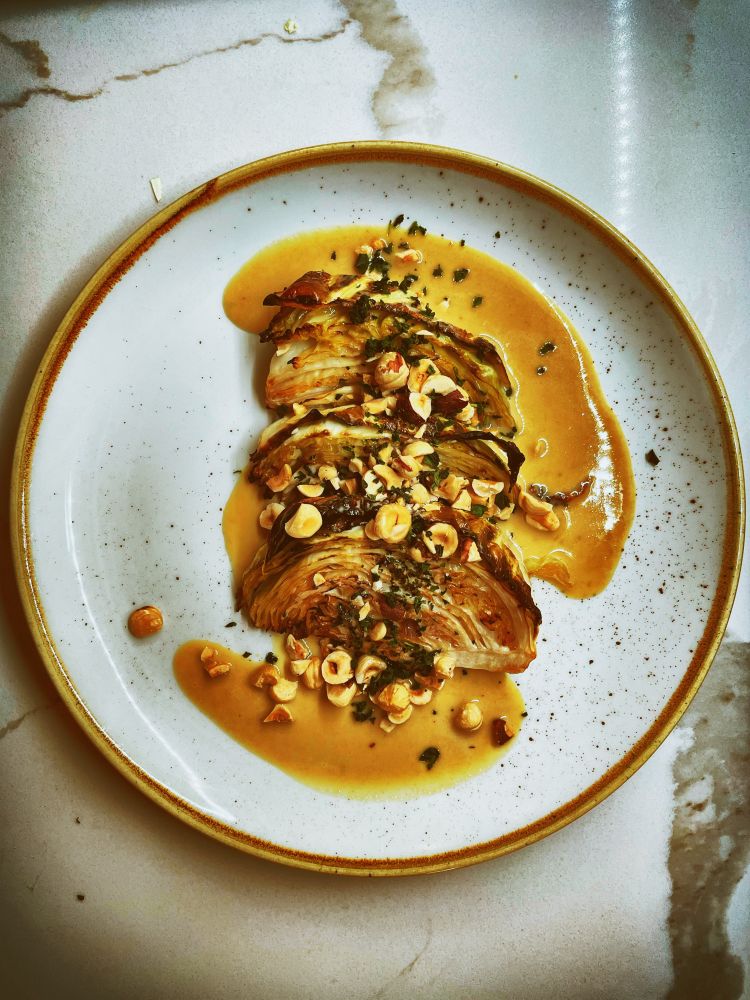 A plate of charred cabbage rests on a glazed ceramic plate, sitting atop a white marble counter. The cabbage is darkly roasted and topped with a golden brown butter tahini sauce, which forms a vague swirl around the cabbage. The cabbage is topped with toasted hazelnuts and fresh minced mint.