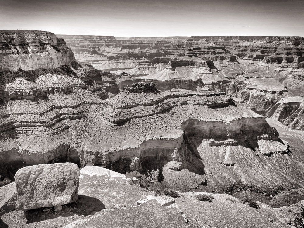 A black and white image of the Grand Canyon. The expanse of the canyon stretches out seemingly endlessly, with various buttes/plateaus fading outward to meet the horizon line. The image was taken at/near noon, and as a result the shadows from the strata starkly contrast against the sunlit areas of the canyon. 