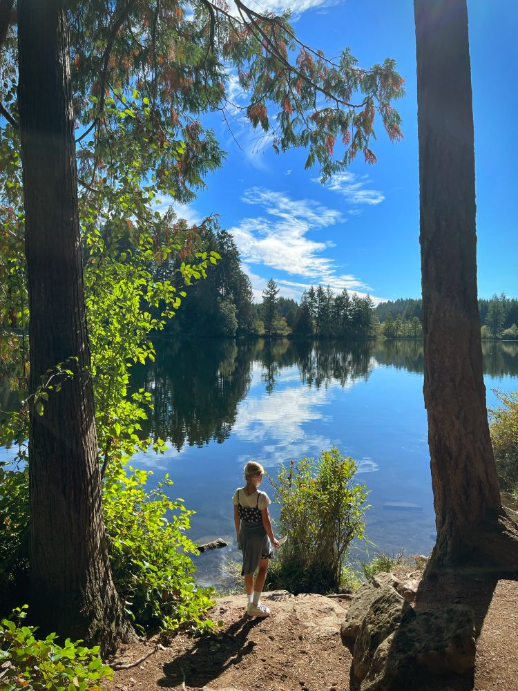 Image of a young girl standing at the edge of a mirrored lake, reflecting the brilliant blue sky and treeline from the opposite bank of the lake. She is framed by large pine trees on either side. 