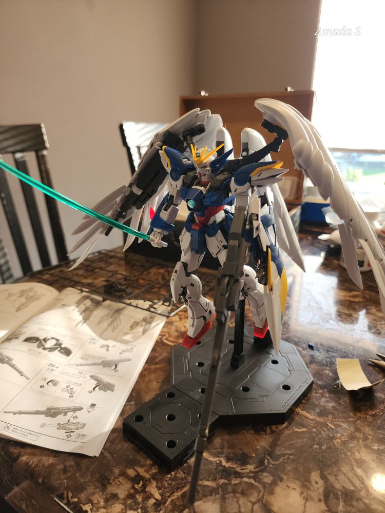 A different angle of the wing zero. A white and blue mecha with red accents holding a beam saber in the right hand and a beam rifle in the left. It has white angelic wings. The instruction booklet is beside it.