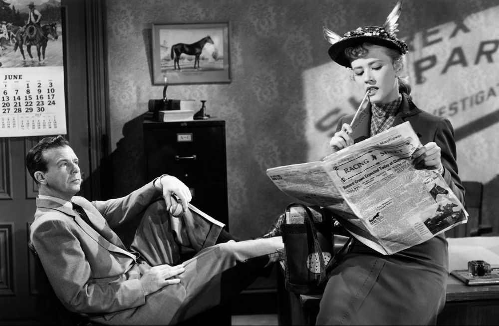 Dick Powell sits in an office chair while Joyce Holden reads the horse racing news in YOU NEVER CAN TELL (1951).