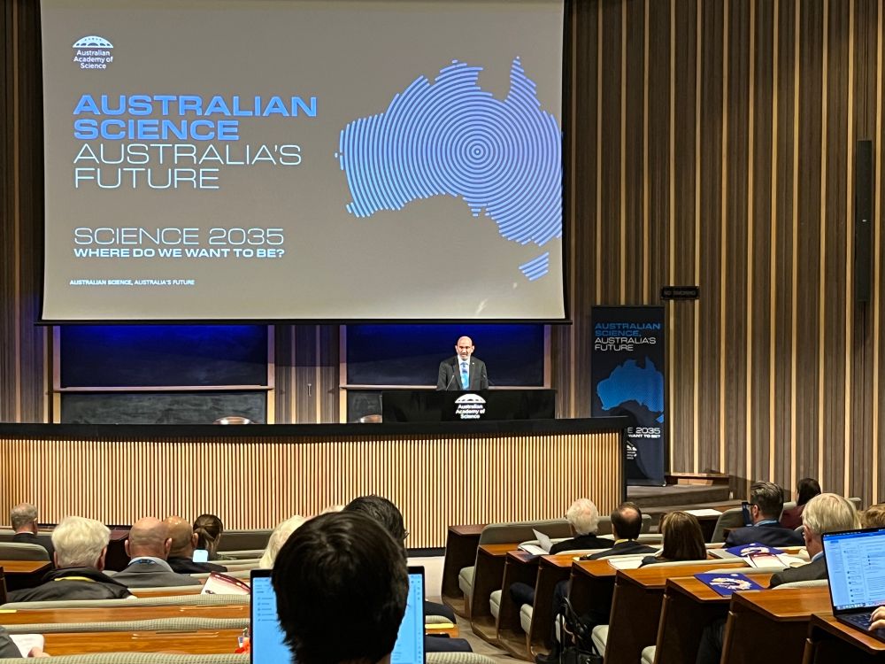 A room with a sloping floor and wooden benches with seated people looking towards the podium with the President at the lectern in front of a screen stating “Australian Science, Australia’s future”.