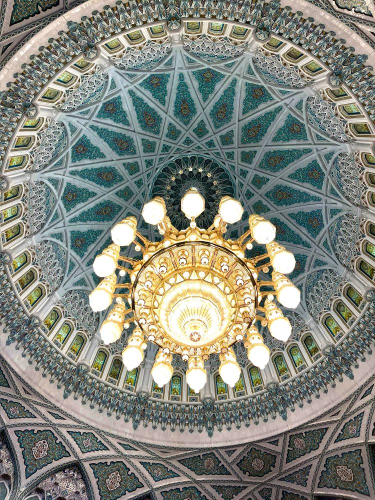 A grand crystal chandelier hanging in the middle of a circular dome with a geometric design of crisscrossing arcs, rimmed with arches.