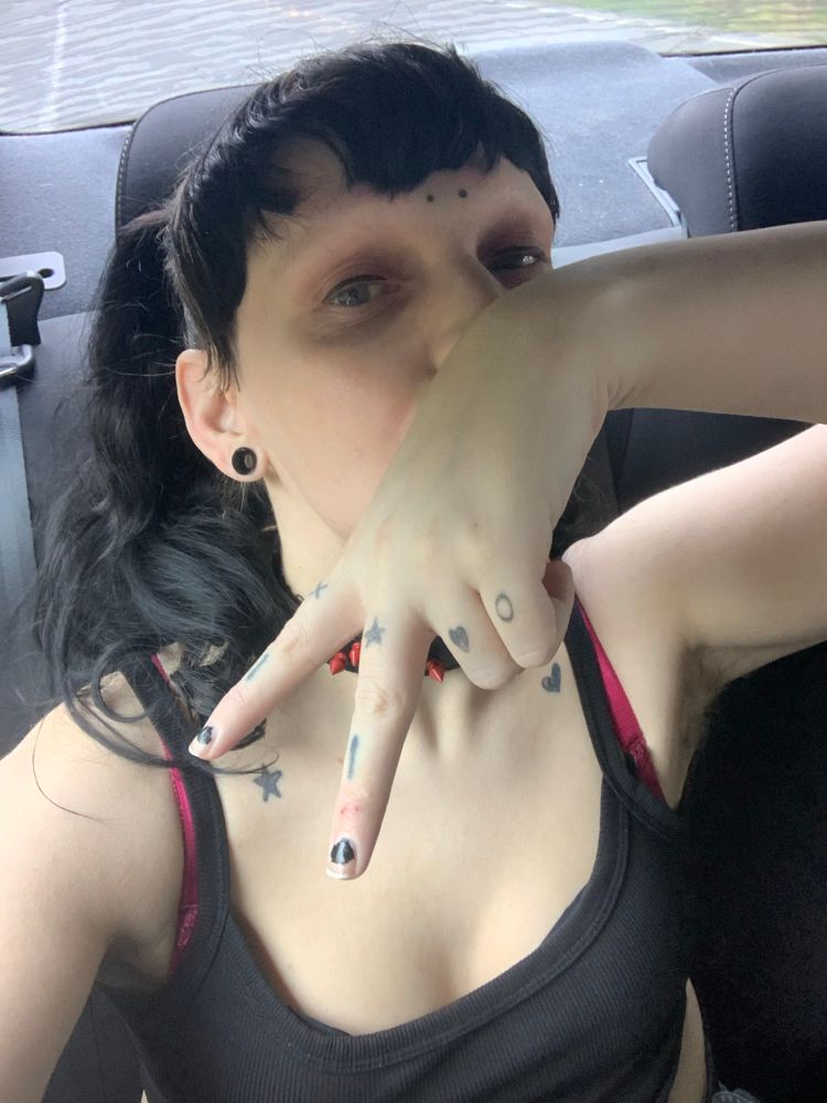 a selfie of zero, it has a black tank top, and black/red spiked collar on. it has black hair and bangs with pigtails. it holding its hand infront of its face with two fingers up 