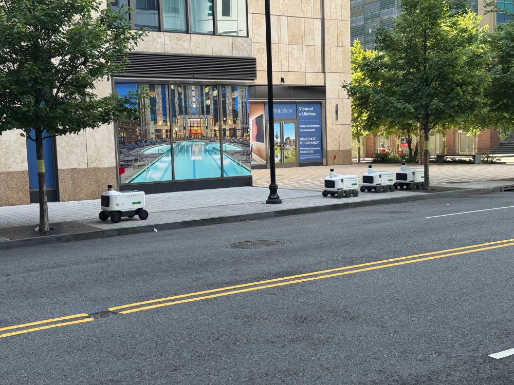 An image of four food delivery robots lined up on a sidewalk. 