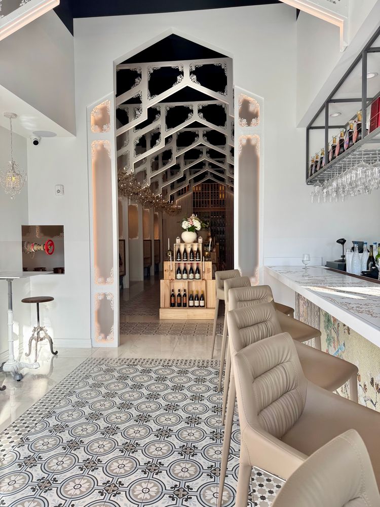 Restaurant interior showing a tile floor patterned in black, white and taupe, leadypast a long white bar counter lined with high taupe chairs. A tall, narrow doorway leads to a darkened dining room beneath stylized white trusses that makes it look like a nave. At the front stands a pale wooden stand showcasing wine bottles.