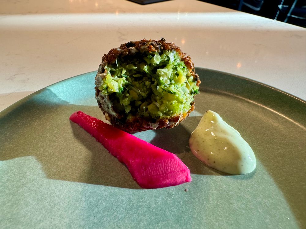 A small globe of falafel in cross-section, with sun catching the varying shades of green in the generously herbed ground-chickpea mixture. The outside looks crackly and deep-brown. On the celadon-green plate sits a vivid magenta spear of pickled turnip and a glossy dab of ivory tzatziki.