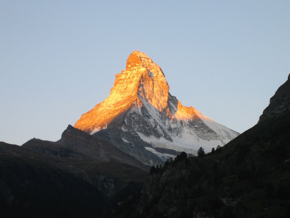 Foto Matterhorn bei Sonnenaufgang.  Markanter, freistehender, spitzer Berg mit oben abknickender Spitze. Ost- und Nordwand von der aufgehenden Sonne orange beleuchtet. Grashänge und bewaldete Bergrücken im Vordergrund im Schatten noch sehr dunkel.
