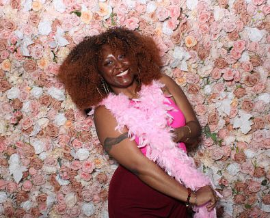 Photo of NuuYawkerr wearing a pink boa against a pink flower wall. 