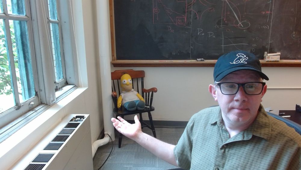 A picture of me sitting at my desk pointing at a wooden chair containing a large stuffed Homer Simpson.  Blackboard with uninterpretable nonsense in the background