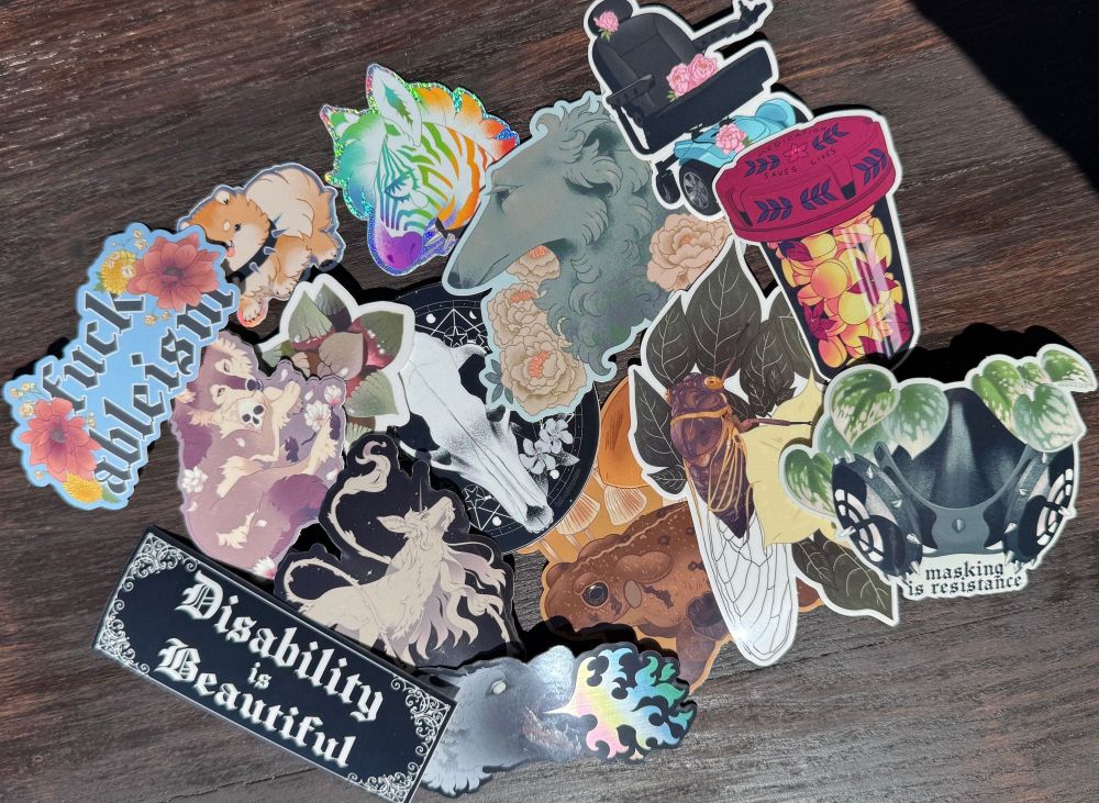 a variety of stickers laid out on a table with various "witchy" animals, florals, and disability themed stickers