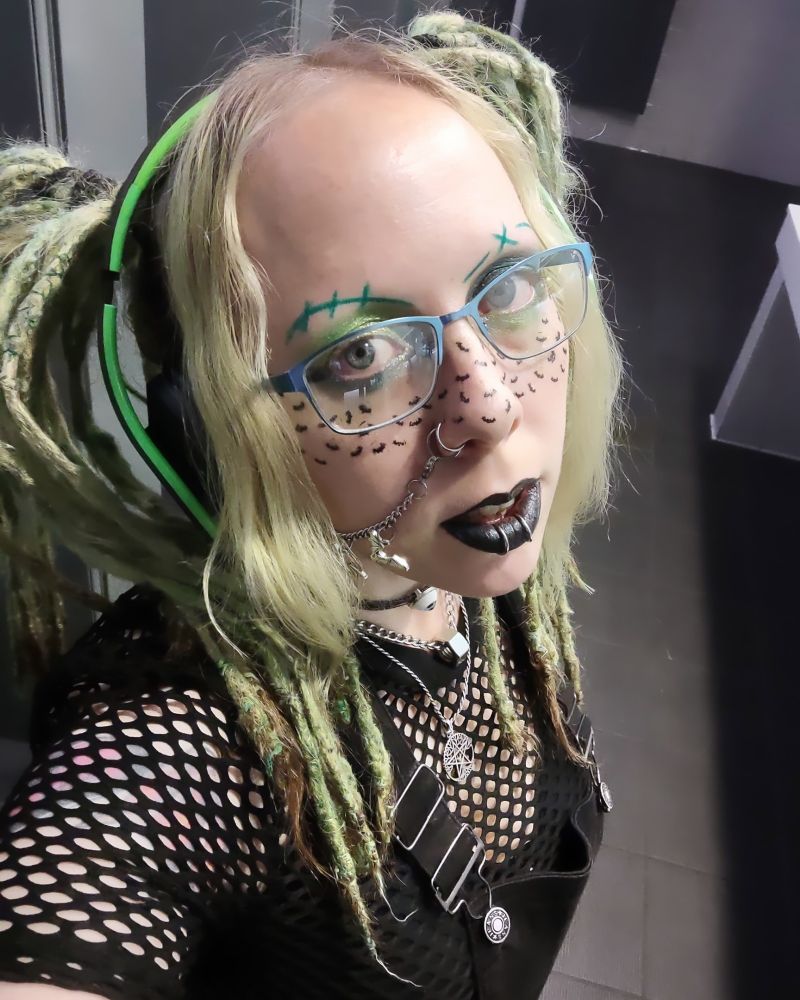 Photo is a goth girl taking a selfie. Her hair is light green, in pigtails. She wears glasses, a fishnet shirt with overalls, over ear headphones, a pentacle necklace, and a thin black choker. She has on vivid makeup with drawn on eyebrows, green eyeshadow, black lipstick, and freckles shaped like black bats.