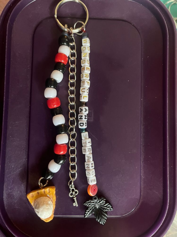 Keychain with three different strands, one of which is black white and red with a pie charm. One is a chain. The third says “wonderful and strange”