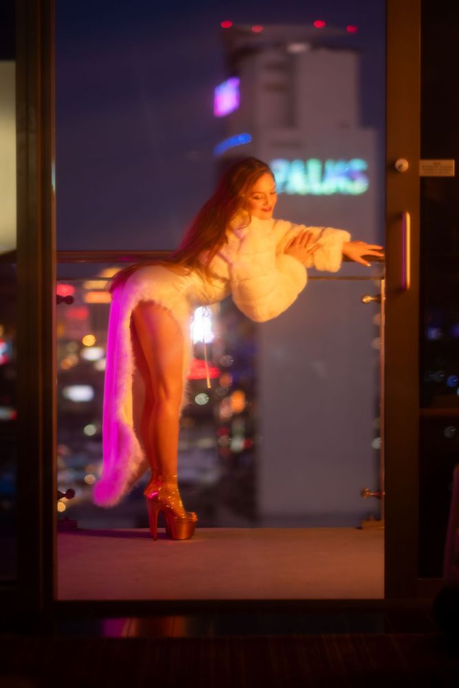 Goldie in white award show dress, leg slit on right towards camera. Multicolor neon lights around.