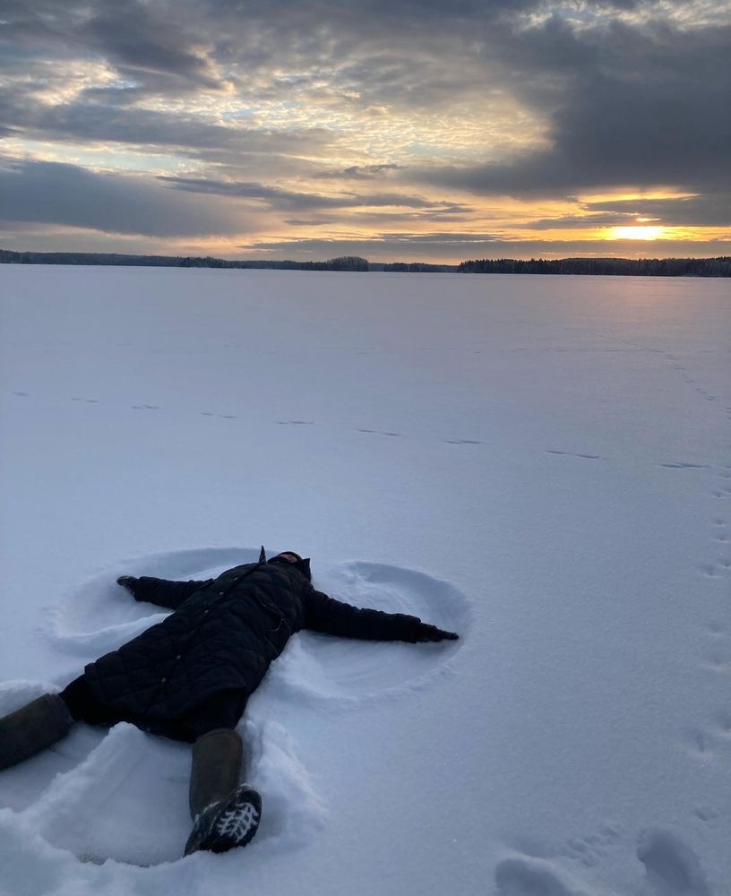 A Person lying on Frozen lake and making an angel pattern to snow. Sun peeking from the clouds in horizon.