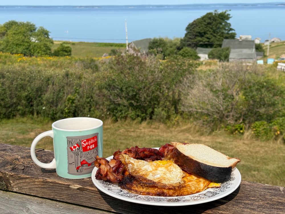 A mug of coffee. A plate with bacon, toast, and eggs fried up in bacon grease. A gorgeous view of Mahone Bay is in the background. 