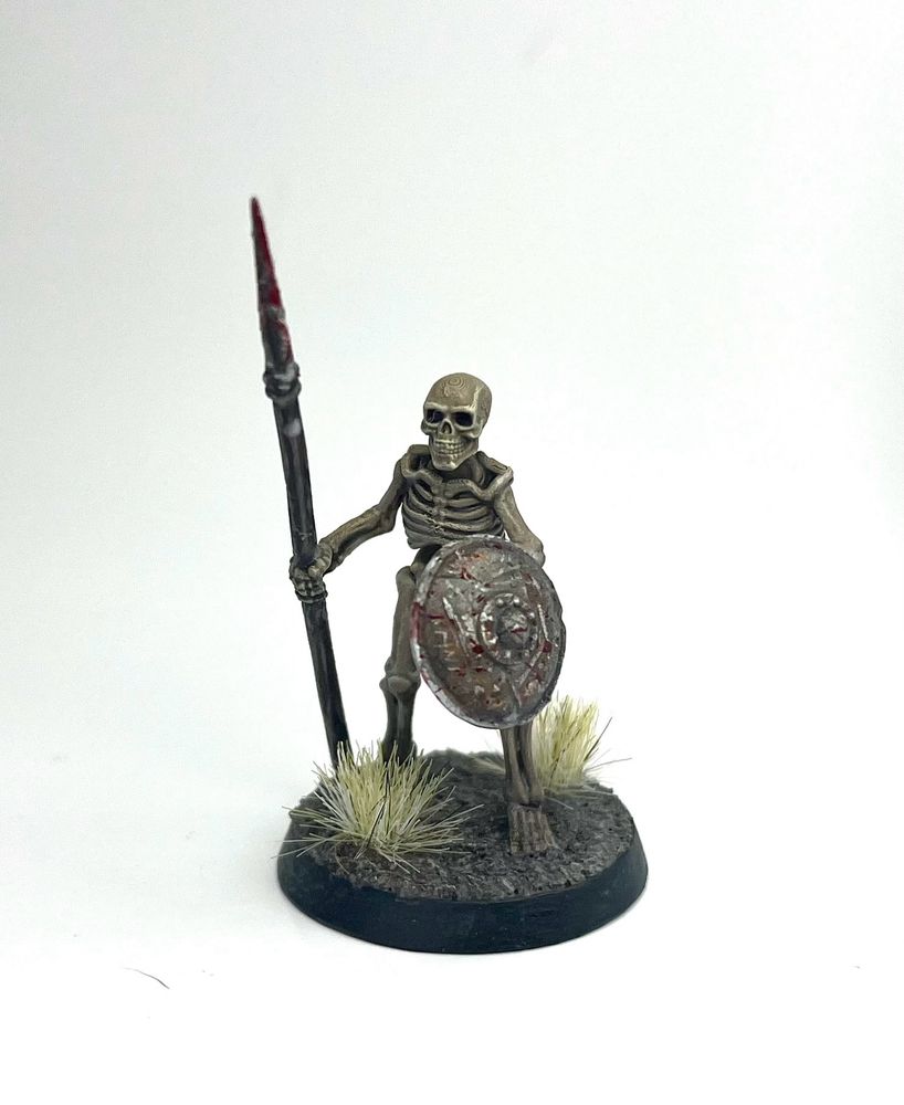 A painted skeleton miniature that is holding a spear and a rusty metal buckler. It is standing amid some dry shrubs growing from the muddy ground.