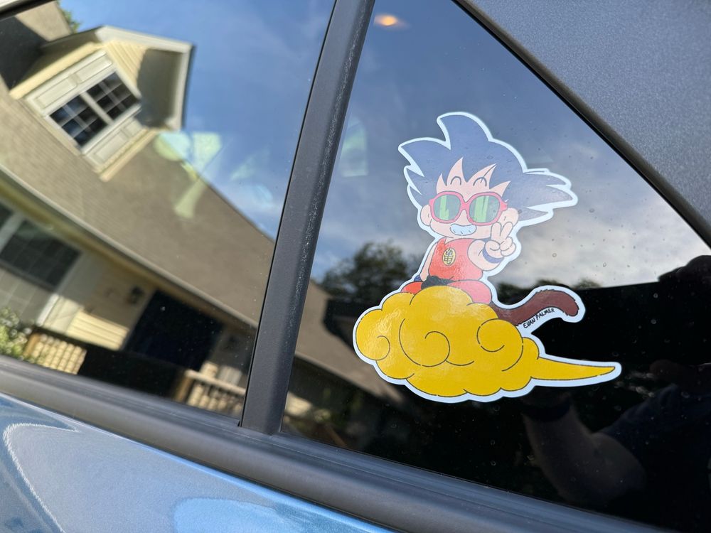 A sticker on a car window. The sticker depicts Goku from Dragon Ball riding his Flying Nimbus while wearing sunglasses. The artist is Evan Palmer. 