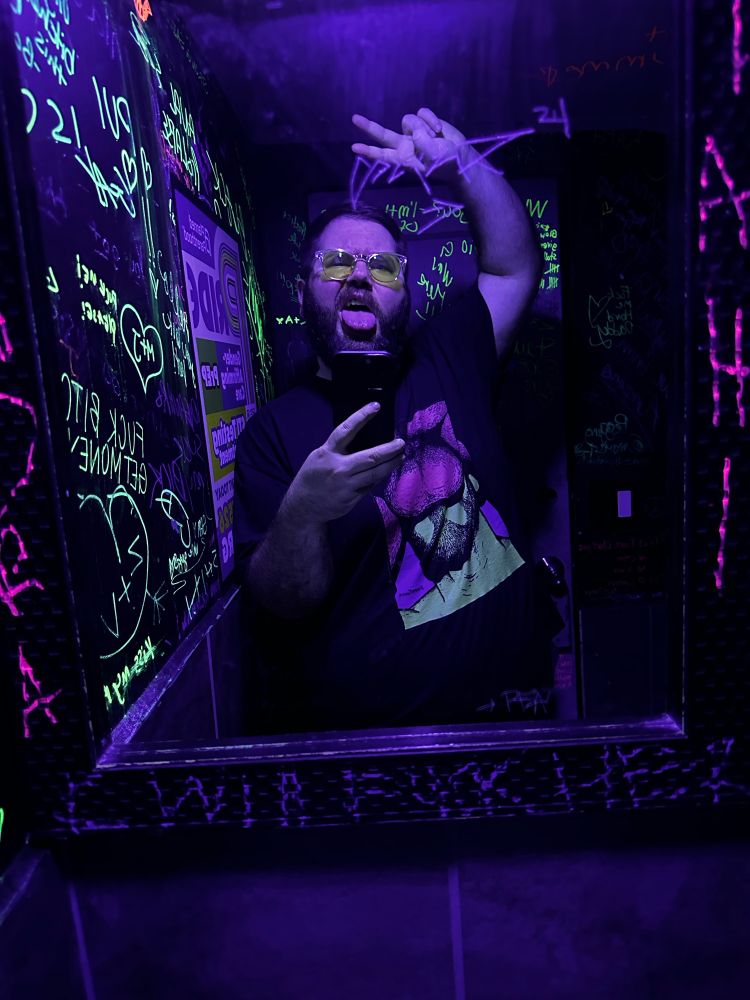White bear guy putting up the peace sign and sticking out his tongue in a neon bathroom wearing a shirt with a man performing oral sex on it.