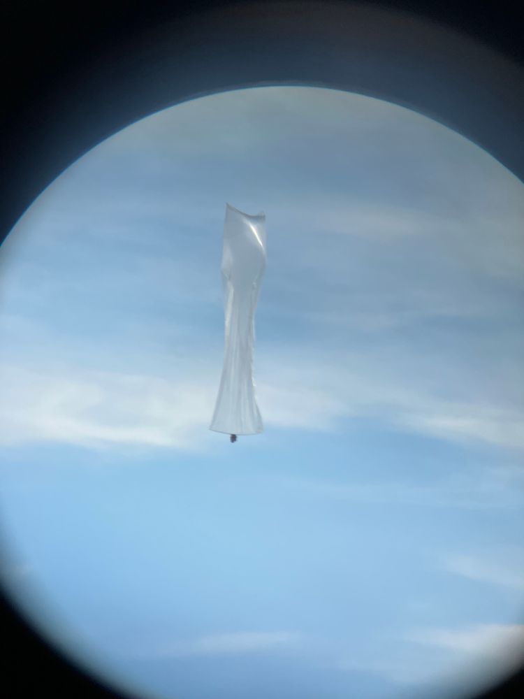 A zoomed in picture of a large translucent balloon with a small dark object attached to the bottom, taken through binoculars.