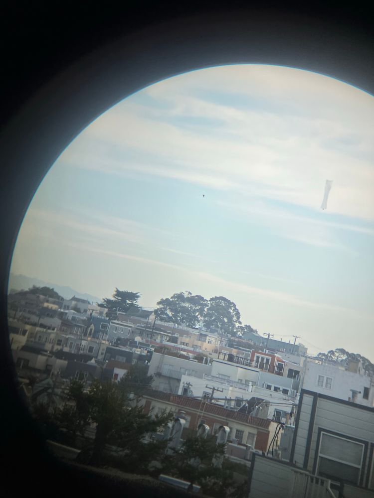 A picture of a tall translucent balloon with a small object attached to the bottom floating over San Francisco. The picture is taken through binoculars.