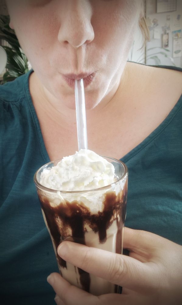 Selfie meines Oberkörpers, zu sehen bin ich von der Nase bis zur Brust. Ich trage ein T-Shirt in Petrol unf trinke einen Eiskaffee durch einen Strohhalm. Am inneren Glasrand läuft Schokosauce nach unten, oben ist ein Sahnehäubchen 