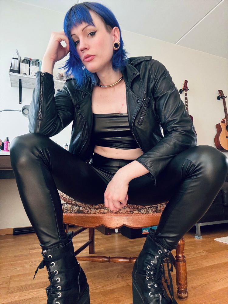 a woman wearing faux leather pants a pair of boots and a black leather jacket her hair is parted behind her ear. she looks kinda intimidating 