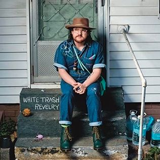 Adeem the Artist is sitting on the backdoor steps of their home. They are wearing a flat brim hat, pink-framed eyeglasses, a blue denim shirt embroidered with a floral design on the right shoulder, blue jeans with rolled up cuff, and green Doc Marten boots. There is a handkerchief extending from the left pocket of the shirt. It is blue, pink, and white in the colors of the transgender pride flag.

The words "White Trash Revelry" are written in chalk on the top step next to where they are sitting.