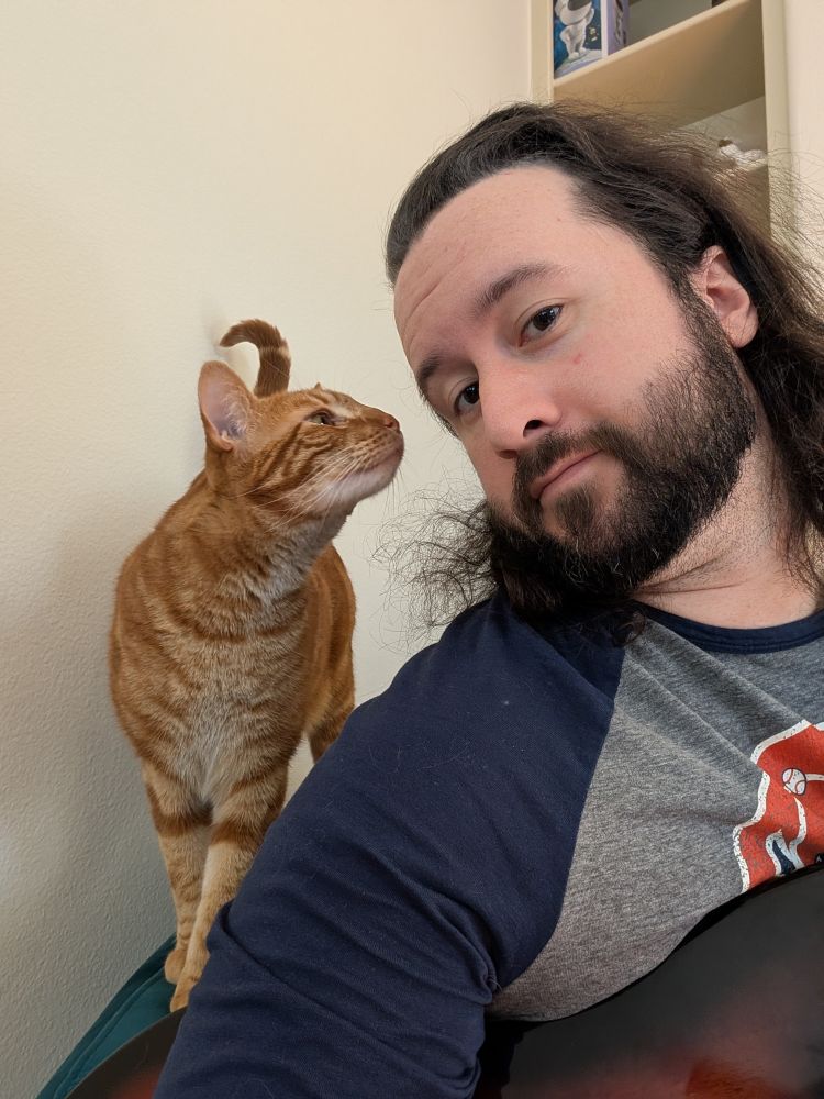 Selfie of me looking at the camera. The body of my bass is partially visible on my lap. A ginger cat is on the arm of the chair next to me, leaning in to sniff my face