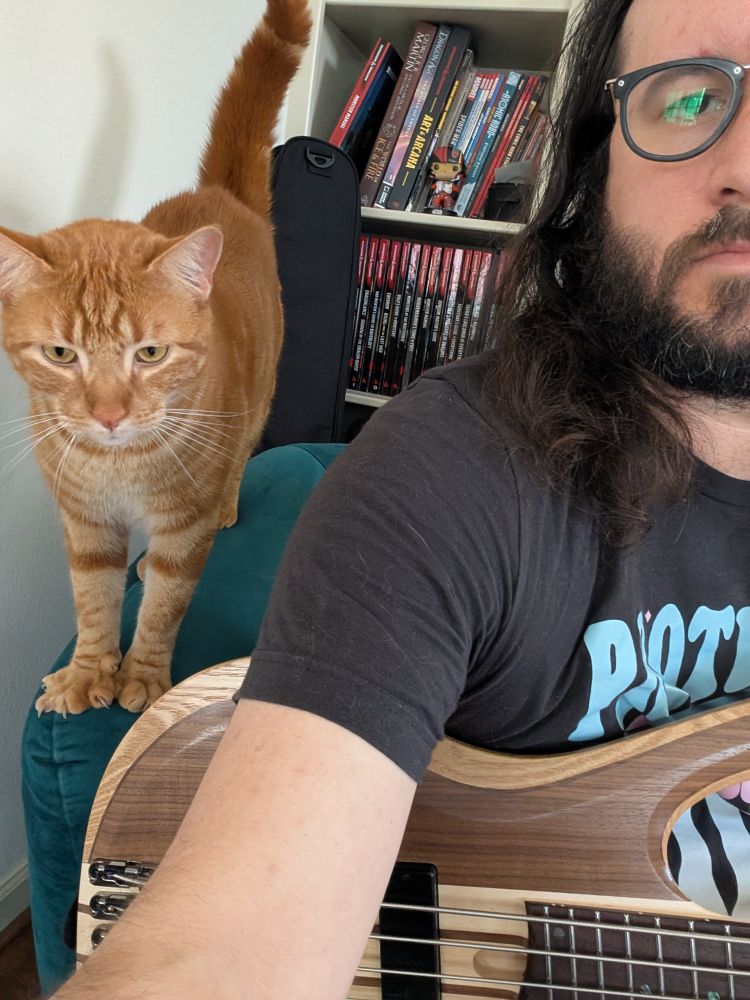 Upper corner of my torso and half my face, bass guitar in my lap, orange cat standing on arm of chair looking at camera