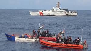 US Coast Guard boats surround a smaller boat, presumably carrying migrants hoping to gain entry to the United States. A white-hulled Coast Guard Cutter lurks in the background