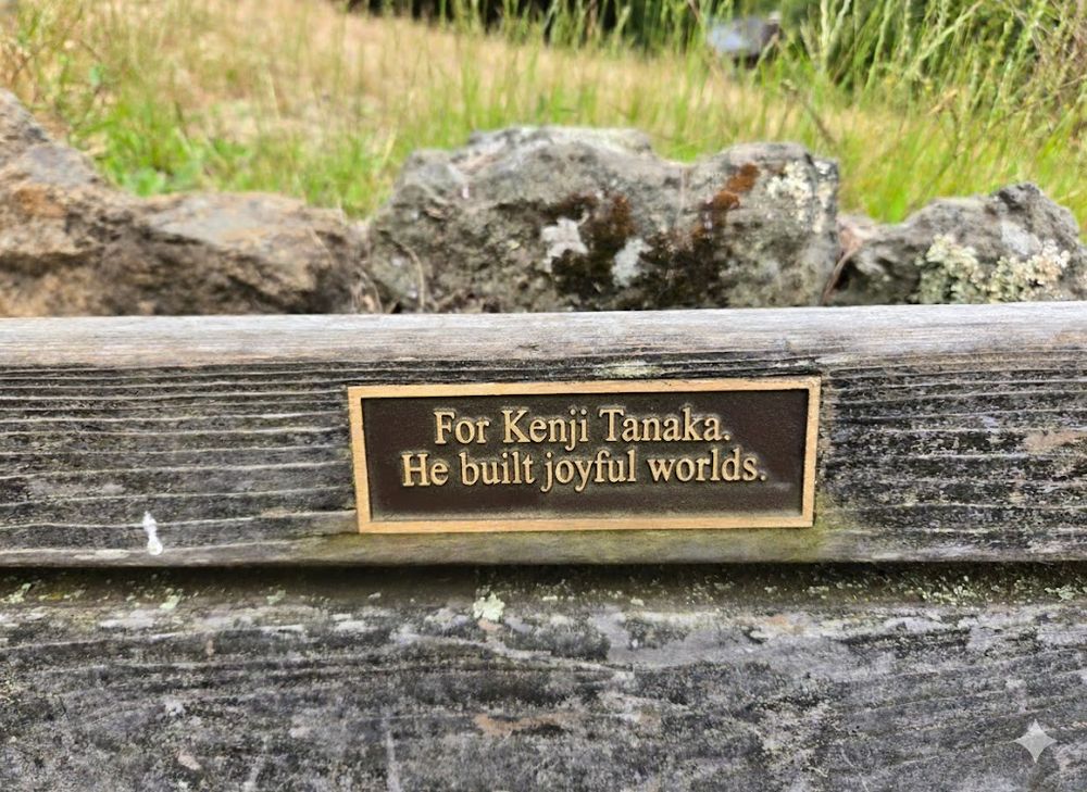 A park bench with a weathered brass dedication plaque that says “For Kenji Tanaka. He built joyful worlds.”  Photo taken by the author in the Berkeley Rose Garden; AI tool used to fictionalize names.