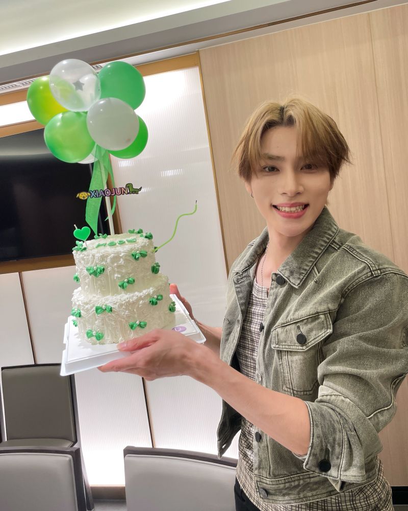 xiaojun in a denim jacket and blond hair, holding a white cake with green ribbons and his name on it, as well as balloons.