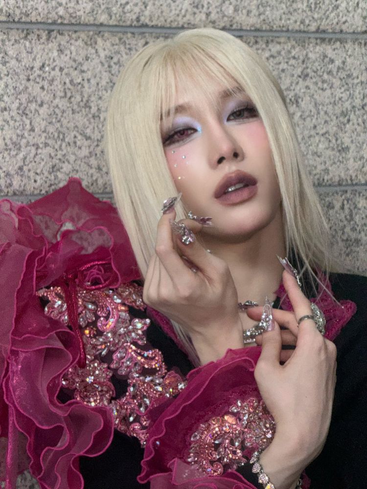 rui with blonde hair and bangs and elaborate makeup and acrylic nails in silver with chains and butterflies. She's in a black outfit full of sequins in pink tones and dark pink ruffles. There are little rhinestones on her cheek.