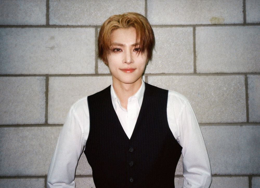 xiaojun looking pretty in a white shirt and blakc vest, with blonde-orange hair, leaning against a brick wall.