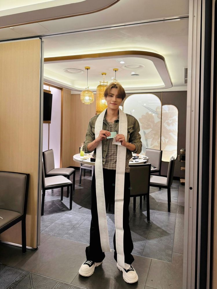 xiaojun standing in a corner of a restaurant with a recipe around his neck that reaches the floor.