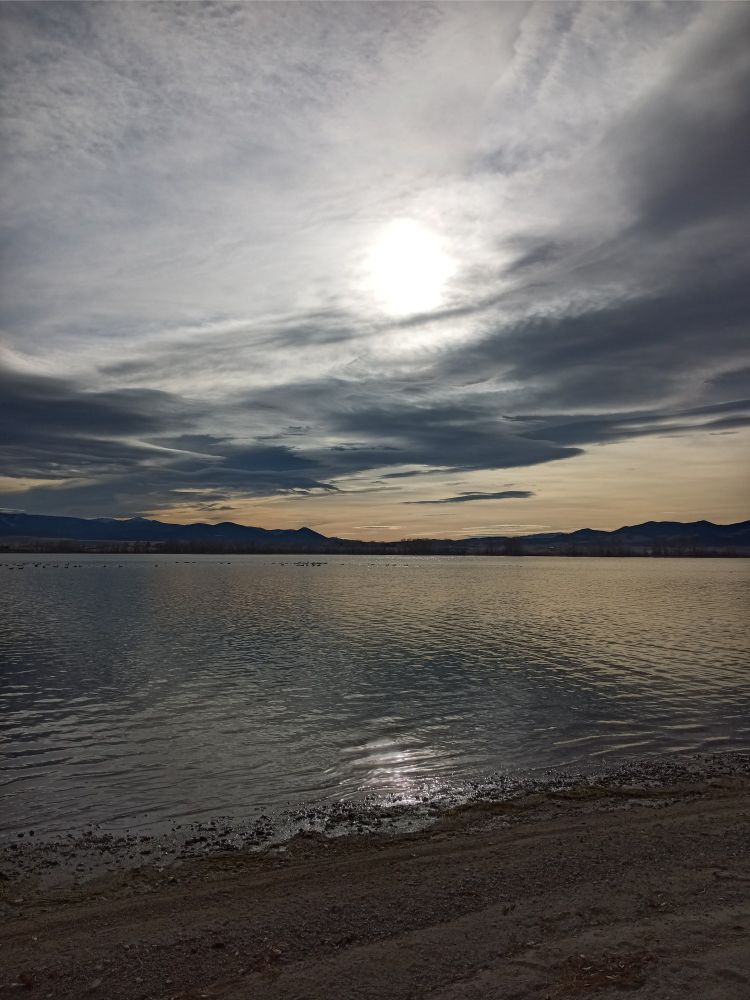 Setting sun pushing through light and heavy grey clouds. Yellow pink light along the horizon. Sun is reflected in a still grey blue lake. Geese are noisy but out of sight.