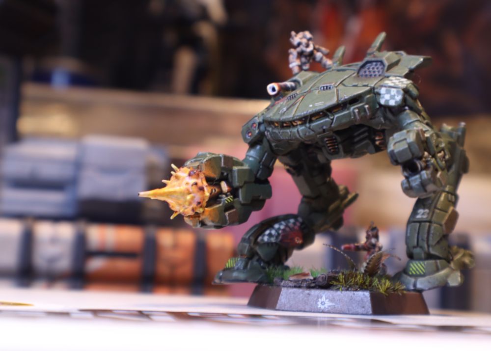 A miniature King Crab, painted in Star League colors, firing its cannon while being swarmed by battle armored troopers.