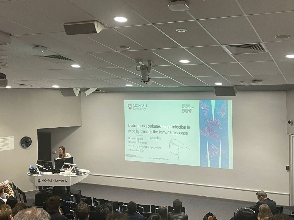 A photo taken from the back of a lecture theatre, showing me at the lectern and the title slide of my talk in the background