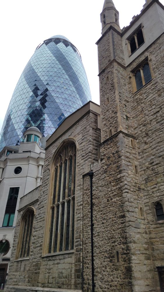 15th century St Andrew Undershaft Church - grey stacked stones - set against 30 St Mary Axe (The Gherkin) - modern, conical all glass building - in London
