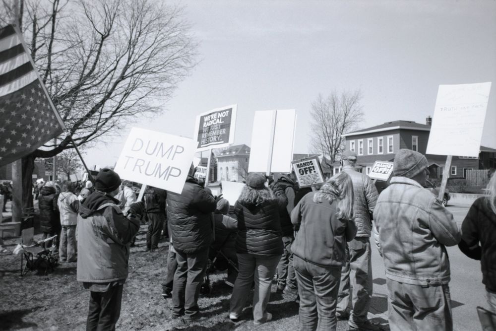 A black and white film photo showcasing some of the signs at the Hands Off protest on Apr 5 2025 in Ludington MI, such as "Dump Trump" and "We're not radical, we just remember history"