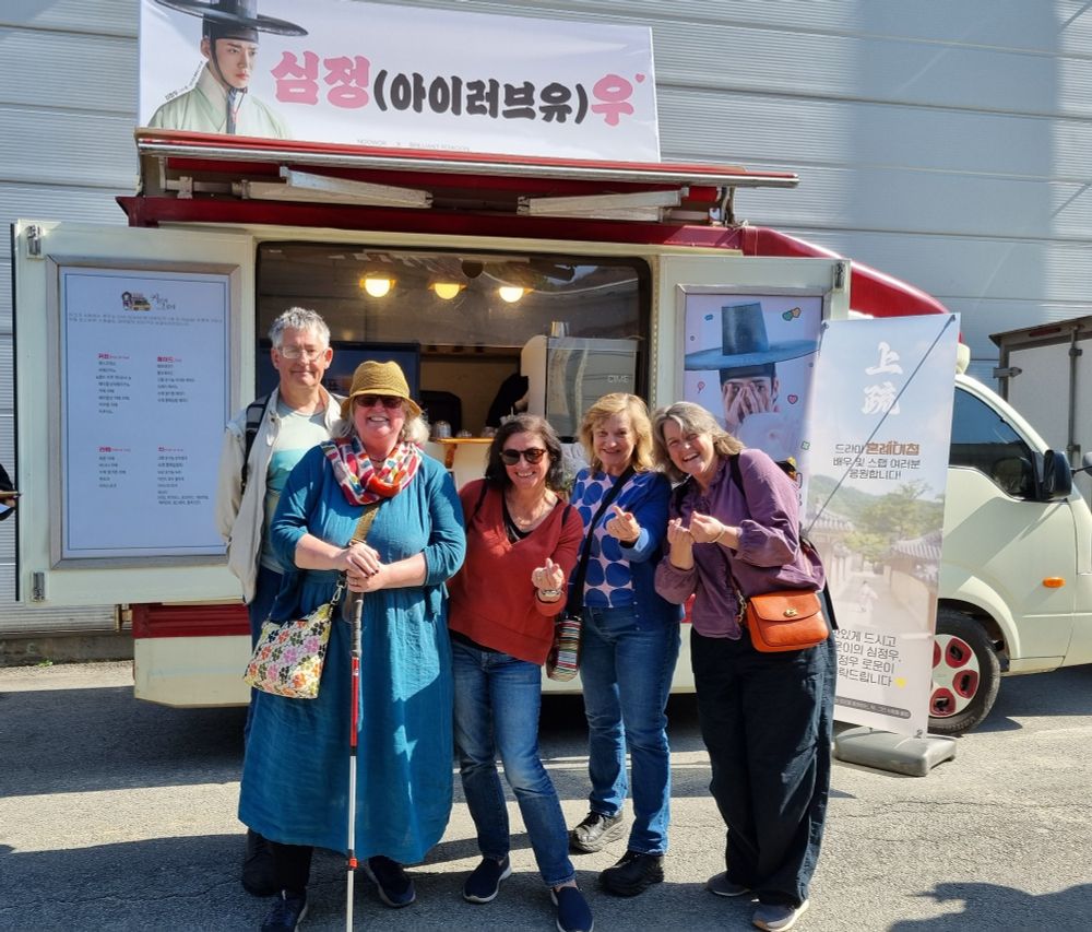 four middle-aged women and one slightly baffled middle-aged man pose with beaming smiles in front of a Rowoon coffee truck on a Korean drama set.