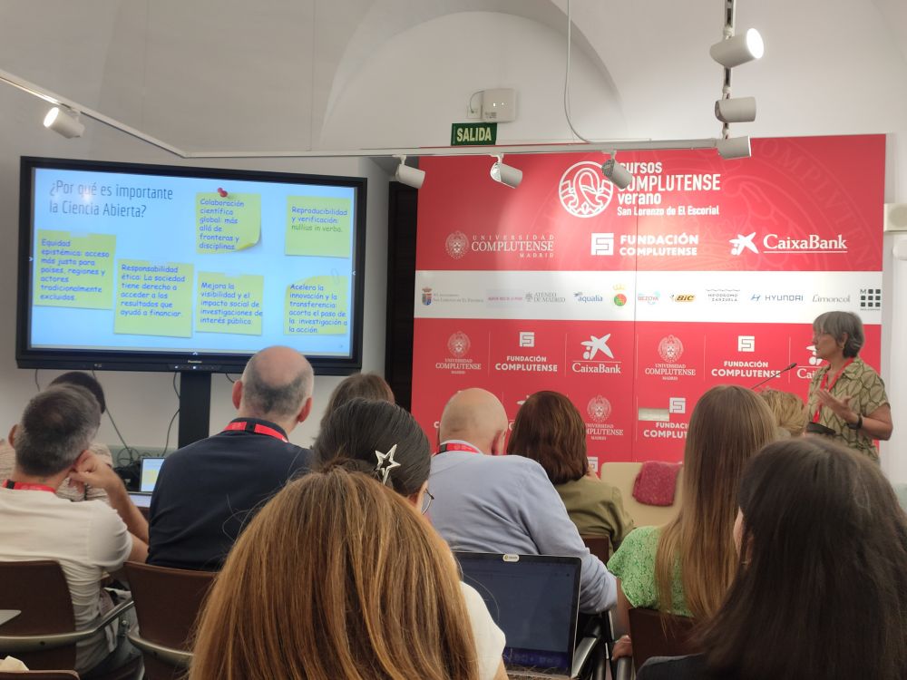 Maite Melero de pie, tras un atril, ante el photocall de los Cursos de Verano Complutenses de El Escorial explica una diapositiva con ideas sobre por qué es importante la ciencia abierta.