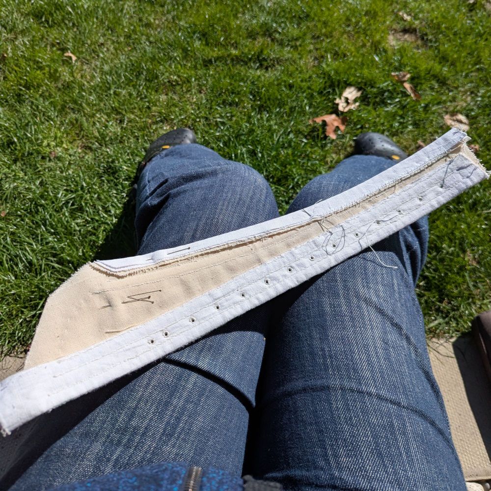 A piece of stays lays on my knees, with seam allowances half stitched down. You can see my legs dressed in jeans and a stretch of sunny grass below my feet.